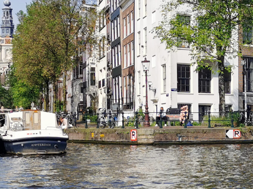 Amsterdam canal in front of white buildings, the front of a white barge on the left, 2 people sitting on a bench looking at the canal.